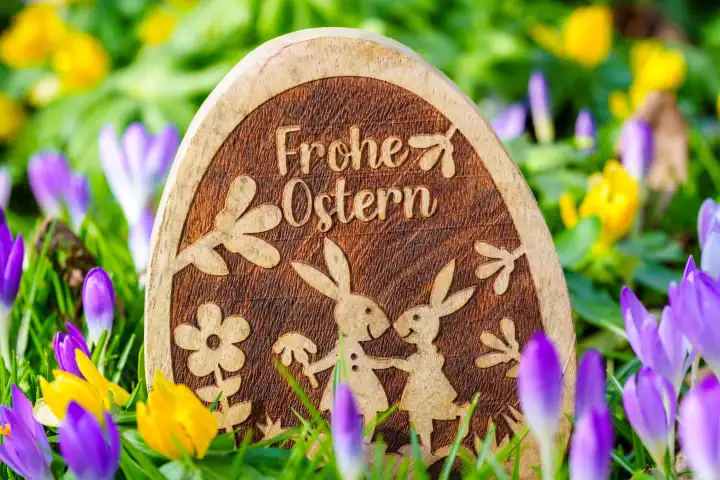 Frohe Ostern! Gruß auf einem Osterei aus Holz das in einer Wiese mit Frühlingsblumen steht. Frohe Ostern Schriftzug.
