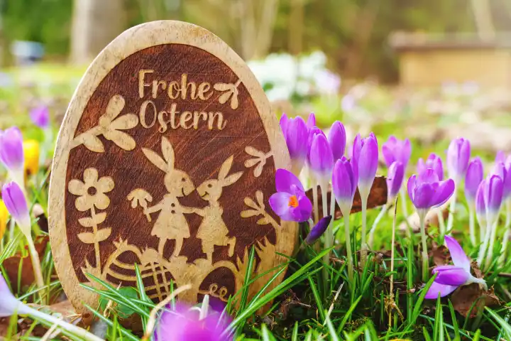 Frohe Ostern! Gruß auf einem Osterei aus Holz das in einer Wiese mit Frühlingsblumen steht. Frohe Ostern Schriftzug.
