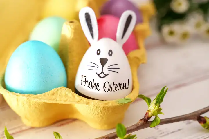 Frohe Ostern! Gruß auf einem Ei mit Osterhasen Ohren und Gesicht in einer Eierschachtel mit bunten Ostereier. FOTOMONTAGE
