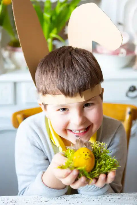 Kind hält ein selbstgebasteltes Osterei mit Hasenohren und Gesicht zu Ostern. Osternest