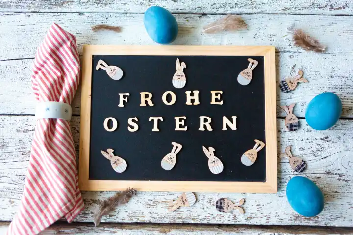 Frohe Ostern, Gruß aus Holzbuchstaben auf einer Kreidetafel mit fröhlichen Osterhasen geschmückt mit blauen Ostereiern