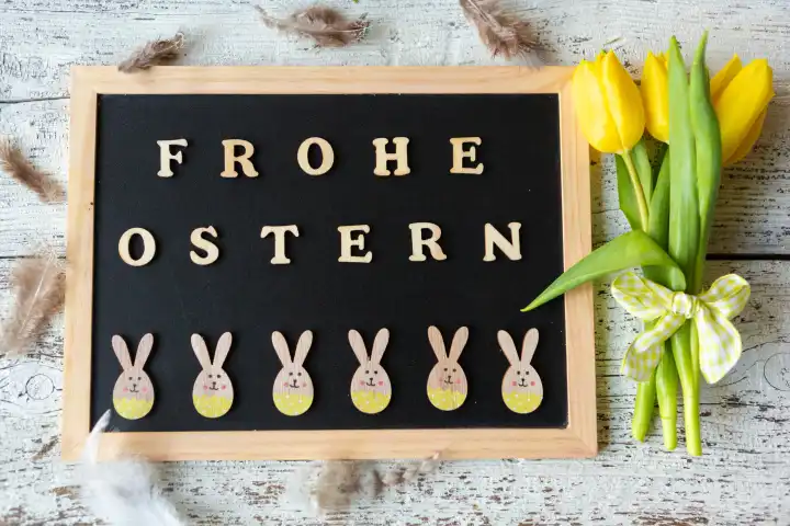 Frohe Ostern, Gruß aus Holzbuchstaben auf einer Kreidetafel mit fröhlichen Osterhasen geschmückt mit gelben Tulpen Blumen