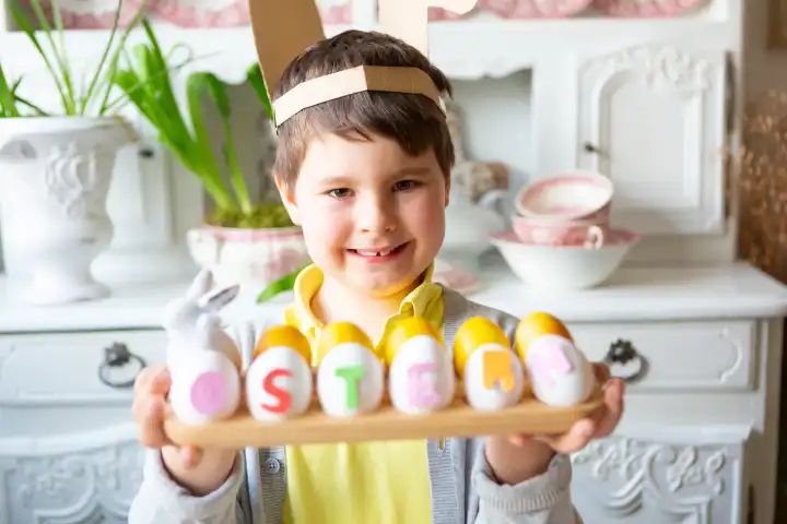 3 March 2024: Little boy making an Easter decoration. Child with bunny ears holds a handmade arrangement with eggs with the inscription: Easter