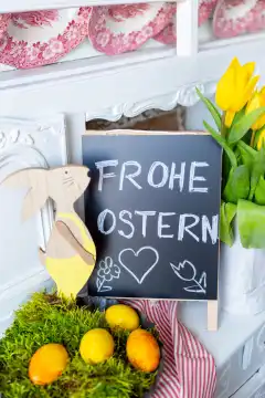 Frohe Ostern, Schriftzug auf einer Kreidetafel. Dekoration zu Ostern mit Osterhase, Tulpen und gelben Ostereiern in einem Nest