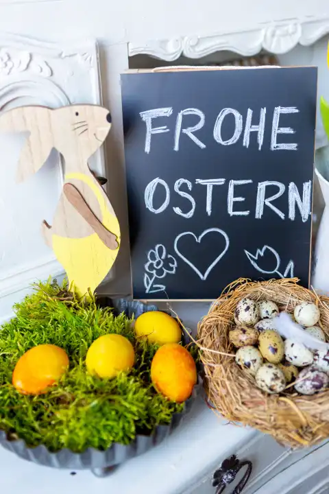 Frohe Ostern, Schriftzug auf einer Kreidetafel. Dekoration zu Ostern mit Osterhase, Tulpen und gelben Ostereiern in einem Nest