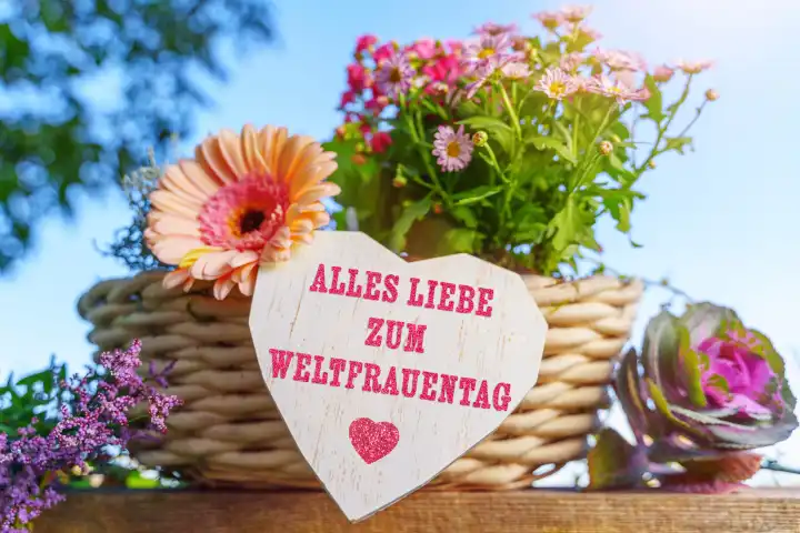Alles Liebe zum Weltfrauentag. Schriftzug auf einem Herz aus Holz im freien bei Sonneschein auf einem Blumengesteck. FOTOMONTAGE