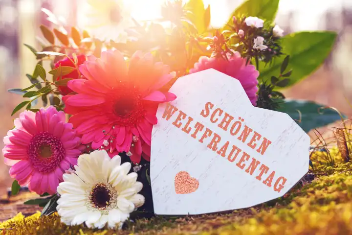 Bavaria, Germany - March 8, 2024: Happy International Women's Day. Greeting for Women's Day on a wooden heart outdoors in the sunshine on a flower arrangement. PHOTOMONTAGE