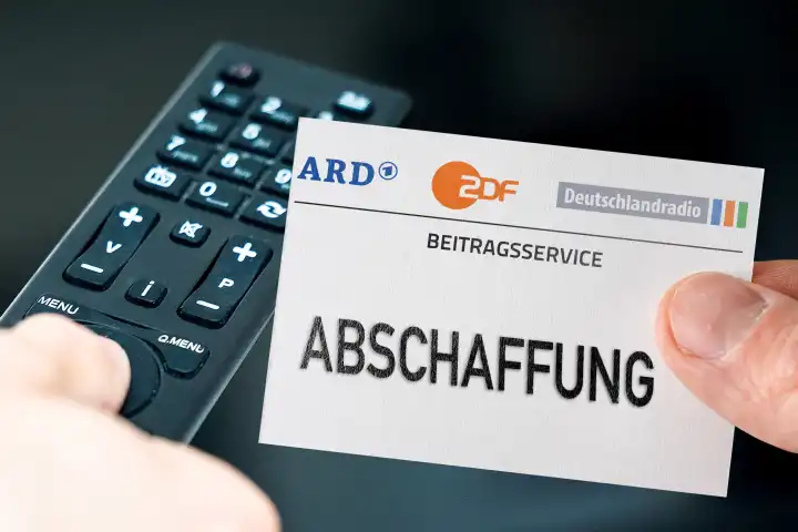GEZ Gebühren Abschaffung Symbolbild. Hand mit ARD, ZDF und Deutschlandradio Logo  vor einer Fernbedienung und einem Fernseher. FOTOMONTAGE