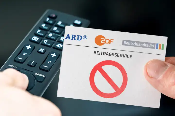 Germany - March 11, 2024: GEZ fees abolition symbol image. Hand with ARD, ZDF and Deutschlandradio logo in front of a remote control and a television. PHOTOMONTAGE