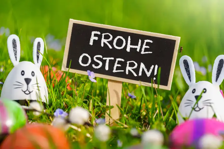 Frohe Ostern! Grüße zu Ostern auf einem Schild in einer Blumenwiese mit bunten Eiern und Eiern mit Hasenohren. FOTOMONTAGE
