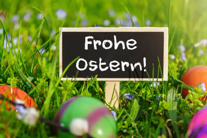 Frohe Ostern! Grüße zu Ostern auf einem Schild in einer Blumenwiese mit bunten Eiern. FOTOMONTAGE 