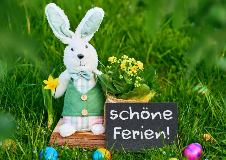 Schöne Ferien! Schriftzug auf einer Tafel neben einem Spielzeug Osterhasen der in einer grünen Wiese mit bunten Ostereiern steht. FOTOMONTAGE