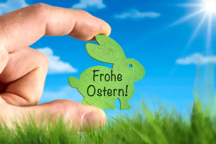 Hand hält Osterhase aus Holz vor einer grünen Wiese und blauem Himmel mit Sonnenschein mit dem Schriftzug: Frohe Ostern! FOTOMONTAGE