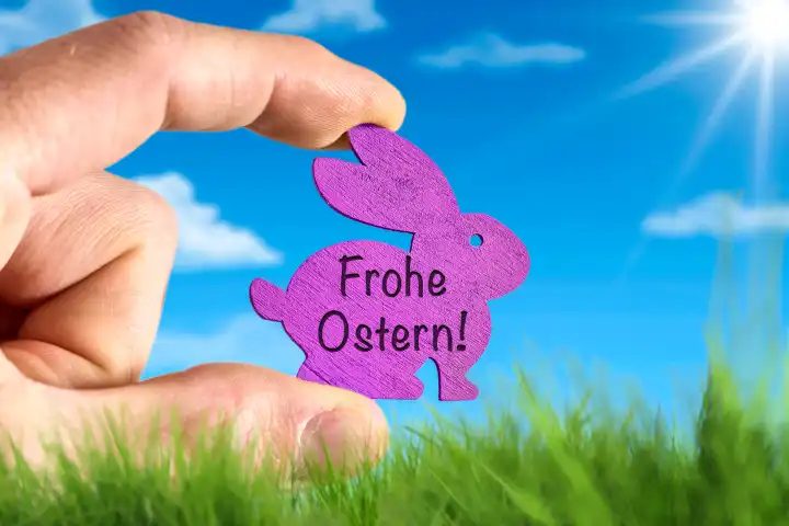 Hand hält Osterhase aus Holz vor einer grünen Wiese und blauem Himmel mit Sonnenschein mit dem Schriftzug: Frohe Ostern! FOTOMONTAGE
