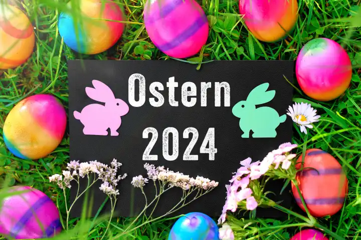 26 March 2024: Easter 2024, lettering on a board in a green meadow with colorful Easter eggs and Easter bunny figures. PHOTOMONTAGE