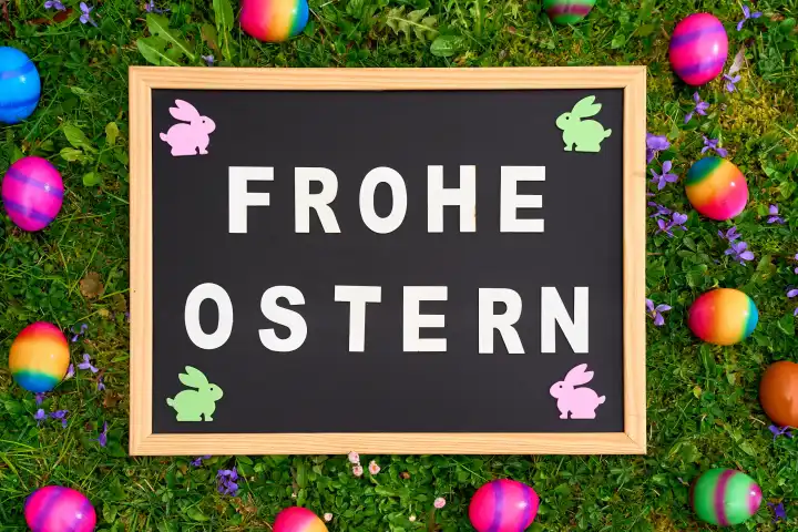 Ostergrüße: FROHE OSTERN! auf einer Tafel in grünem Gras mit bunten Ostereiern