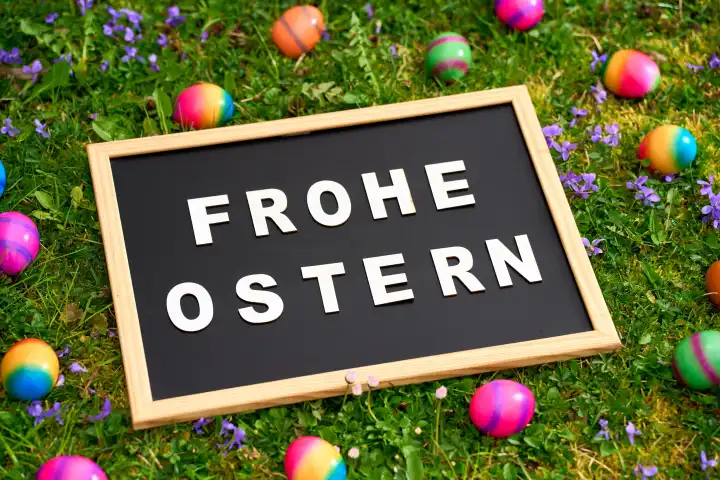 Ostergrüße: FROHE OSTERN! auf einer Tafel in grünem Gras mit bunten Ostereiern