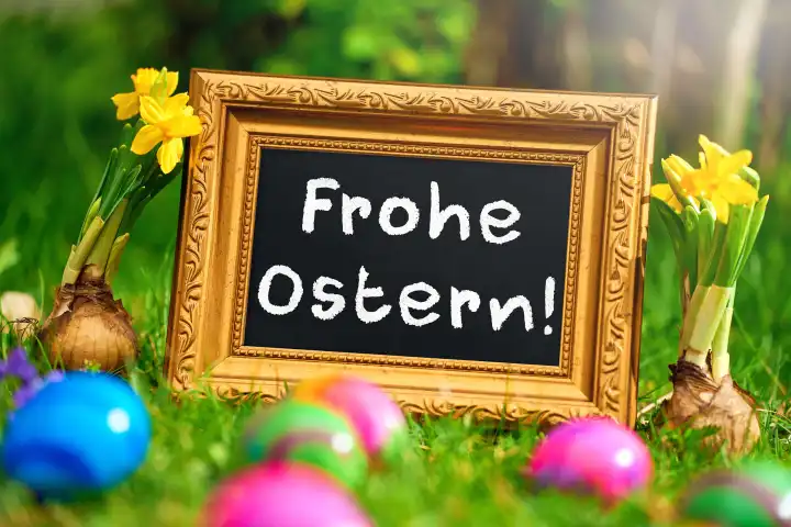 Frohe Ostern! Schriftzug auf einer Tafel mit goldenem Rahmen in einer grünen Wiese mit Frühlingsblumen und bunten Ostereiern. FOTOMONTAGE