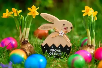 Osterhase aus Holz in einer grünen Wiese mit bunten Eiern und dem Schriftzug: Frohe Ostern