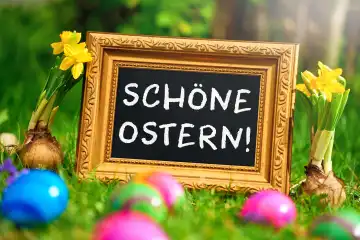 Schöne Ostern! Schriftzug auf einer Tafel mit goldenem Rahmen in einer grünen Wiese mit Frühlingsblumen und bunten Ostereiern. FOTOMONTAGE