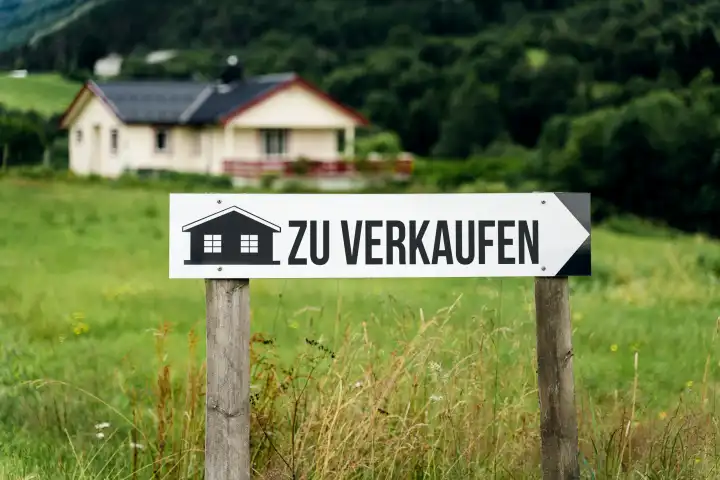 Schild in einer Wiese vor einem Landhaus mit Aufschrift: Zu Verkaufen. Immobilien Konzept FOTOMONTAGE