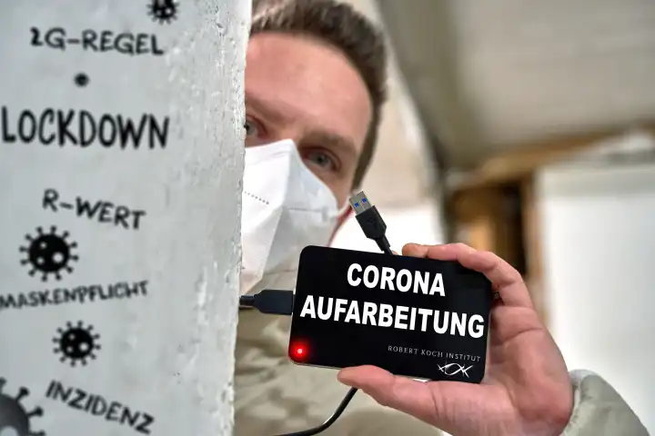 Augsburg, Bavaria, Germany - March 28, 2024: Topic image on the redacted Robert Koch Institute protocols on Covid-19 risk assessments in Germany. A man with FFP2 mask holds a hard disk in his hand with the inscription: Corona Aufarbeitung and RKI logo.  PHOTOMONTAGE
