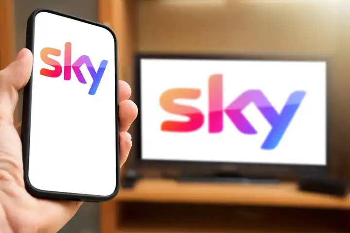 6 April 2024: Hand holding smartphone in front of TV screen with the Sky pay TV and streaming provider logo. PHOTOMONTAGE