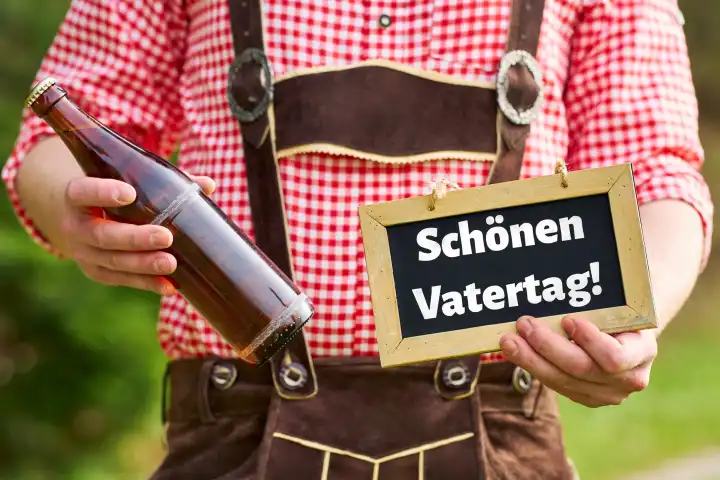 Junger Mann in bayerischer Tracht hält eine Flasche Bier und ein Schild mit dem Gruß: Schönen Vatertag. FOTOMONTAGE