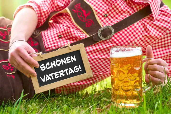 Bavaria, Germany - April 7, 2024: Young man in lederhosen lying in a meadow with a pint of beer and a sign with the greeting: Happy Father's Day! PHOTOMONTAGE