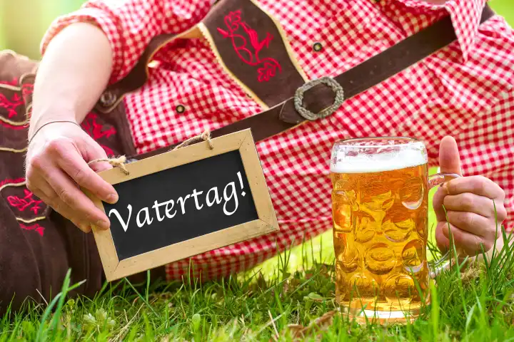 Vatertag! Schriftzug auf einer Tafel die ein Mann in bayerischer Lederhose und Trachtenhemd zusammen mit einem Maßkrug Bier hält. FOTOMONTAGE