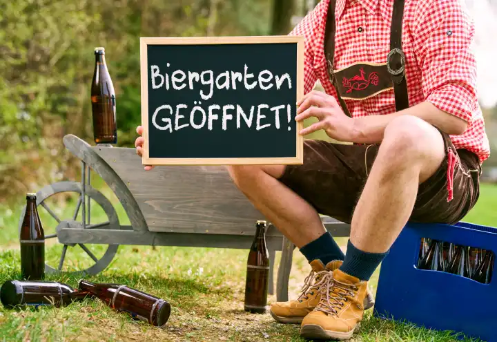 Biergarten geöffnet! Schriftzug auf einem Schild das ein Mann in bayerischer Tracht vor einen Bollerwagen mit Bierkiste und Bierflaschen hält. Biergarten Saison. FOTOMONTAGE
