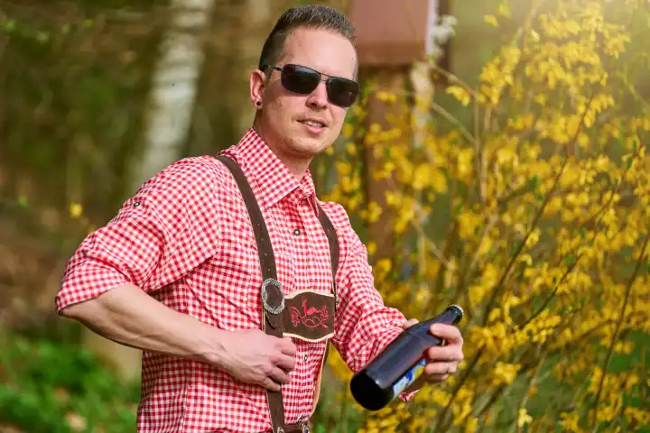 Mann in Lederhose und Trachtenhemd trägt eine Sonnenbrille und hält eine Flasche Bier. Feiern in Bayern Konzept
