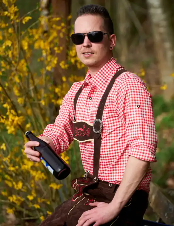 Mann in Lederhose und Trachtenhemd trägt eine Sonnenbrille und hält eine Flasche Bier. Feiern in Bayern Konzept