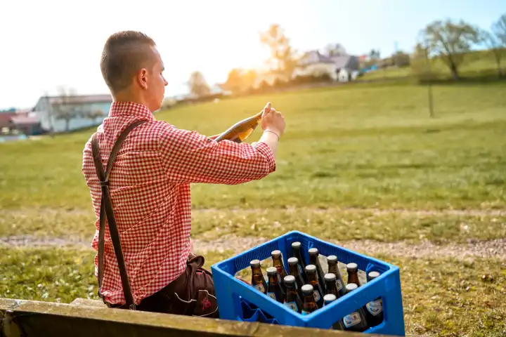 Junger Mann in Lederhose und Trachtenhemd mit einer Kiste Bier auf einer Parkbank. Symbolfoto Volksfest und Vatertag 