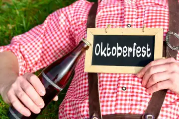 Ein Mann in bayerischer Tracht und Lederhose liegt in einer Wiese an einem Sonnigen Tag mit einer Bierflasche und hält ein Schild mit Aufschrift: Oktoberfest. FOTOMONTAGE