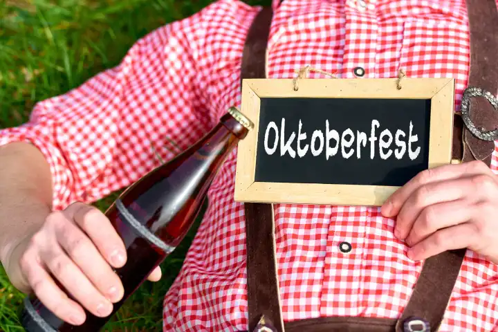 9 April 2024: A man in traditional Bavarian costume and lederhosen is lying in a meadow on a sunny day with a bottle of beer and holding a sign that reads: Oktoberfest. PHOTOMONTAGE