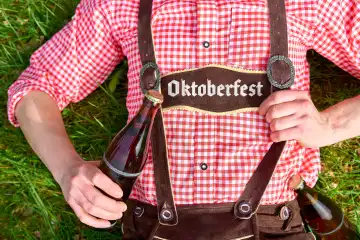 9 April 2024: A man in traditional Bavarian costume and lederhosen lies in a meadow on a sunny day with a beer bottle and the words: Oktoberfest. PHOTOMONTAGE