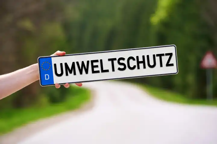 15 April 2024: Hand holding a license plate in front of a country road in green nature with the inscription: Environmental protection. PHOTOMONTAGE