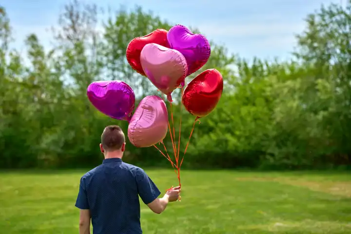 Junger Mann läuft mit roten Helium Luftballons in Herzform über eine Wiese. Symbolbild Liebe und Romantik