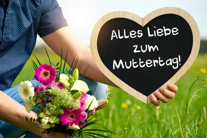 Alles Liebe zum Muttertag! Gruß zum Muttertag, auf einer Tafel in Herzform die ein Mann auf einer Wiese mit einem Blumenstrauß hält. FOTOMONTAGE