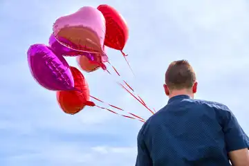 14 April 2024: Young man with red helium balloons in the shape of a heart against a blue sky in sunshine. Symbolic image of love and romance