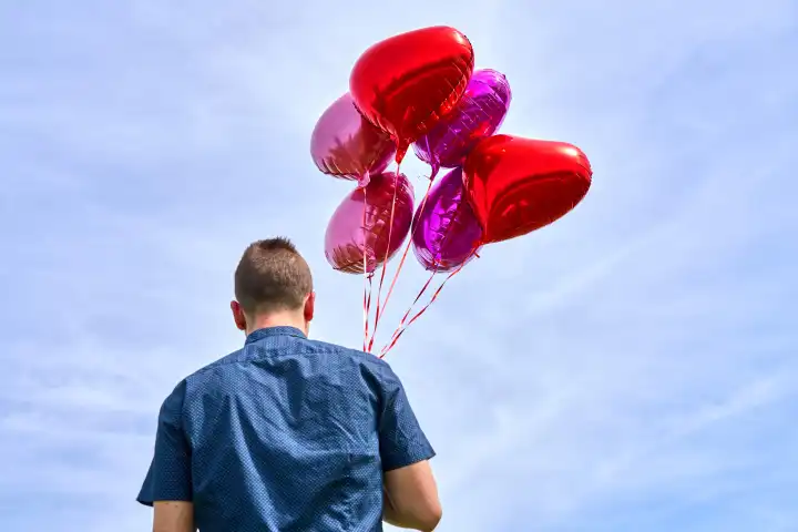 Junger Mann mit roten Helium Luftballons in Herzform vor blauem Himmel bei Sonnenschein. Symbolbild Liebe und Romantik