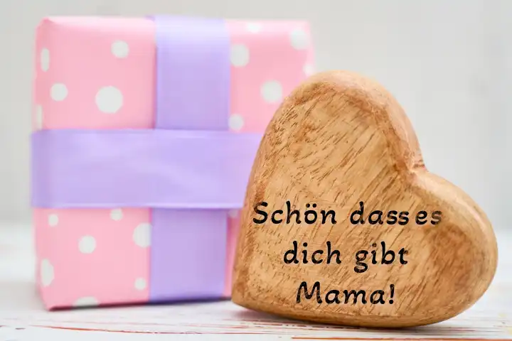 Schön dass es dich gibt Mama! Spruch zum Muttertag, auf einem Herz aus Holz neben einem Geschenk. FOTOMONTAGE