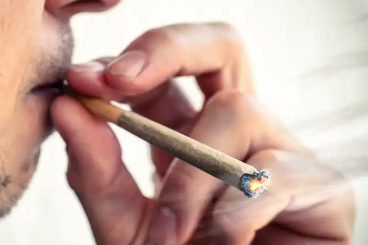 Mann raucht bzw. kifft einen Joint aus Cannabis bzw. THC in der Öffentlichkeit. Nahaufnahme beim Kiffen nach der Cannabis-Legalisierung. FOTOMONTAGE