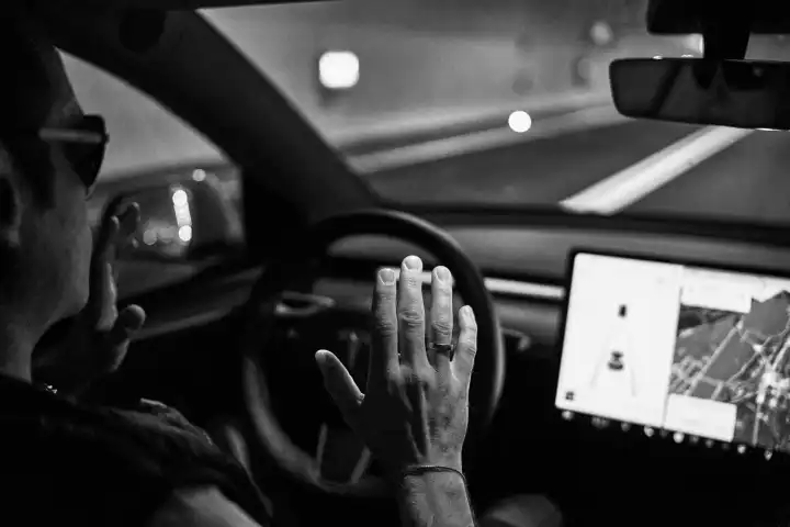 Symbolbild Unfall durch Autonomes Fahren, Ein Mann fährt mit einem Tesla E-Auto mit Autopilot. Schwarz Weiß Foto, hände nicht am Lenkrad