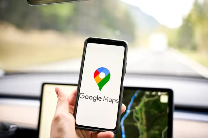 Google Maps Navigation Logo auf einem Smartphone in einem Auto vor einem Navi bzw. Navigationsgerät 