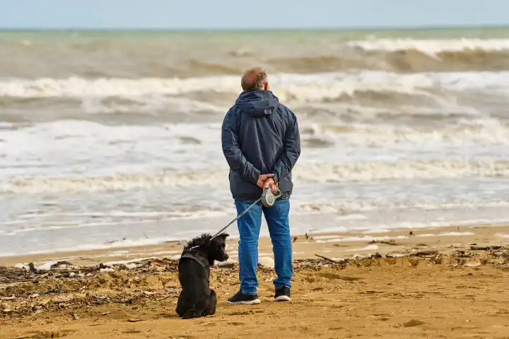 Alter Mann schaut auf das Meer hinaus während er mit seinem Hund an der Leine durch den Sandstrand spazieren geht