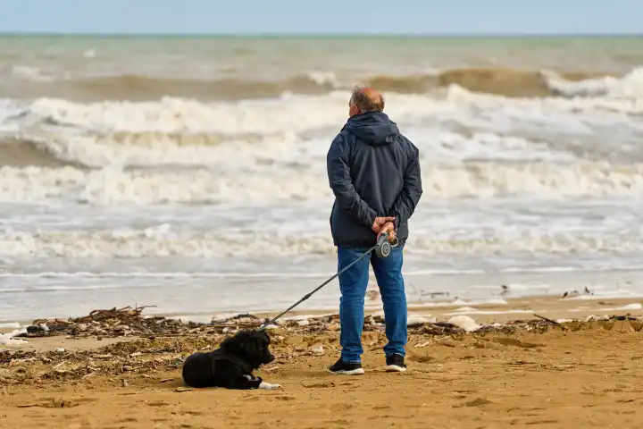 Lido di Jesolo, Italy - 2 May 2024: Old man looking out to sea while walking his dog on a leash along the sandy beach
