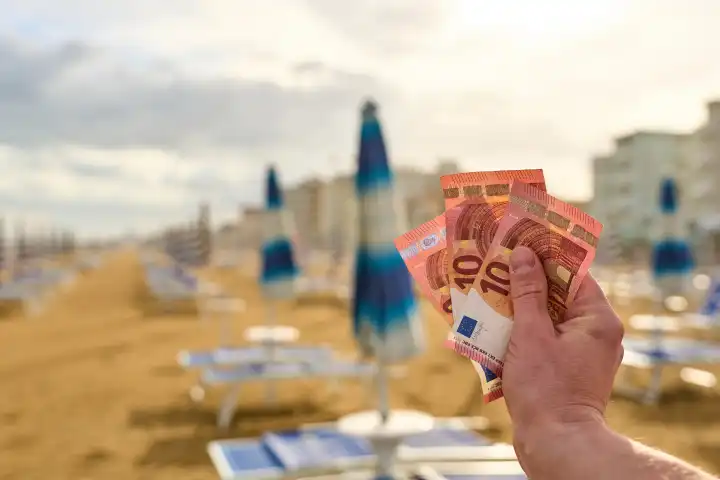 Lido di Jesolo, Italy - 2 May 2024: A man holds euro banknotes in front of sun loungers and parasols on the sandy beach. Vacation money and additional vacation costs concept