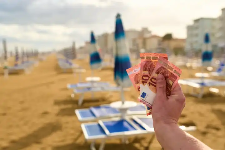 Lido di Jesolo, Italy - 2 May 2024: A man holds euro banknotes in front of sun loungers and parasols on the sandy beach. Vacation money and additional vacation costs concept